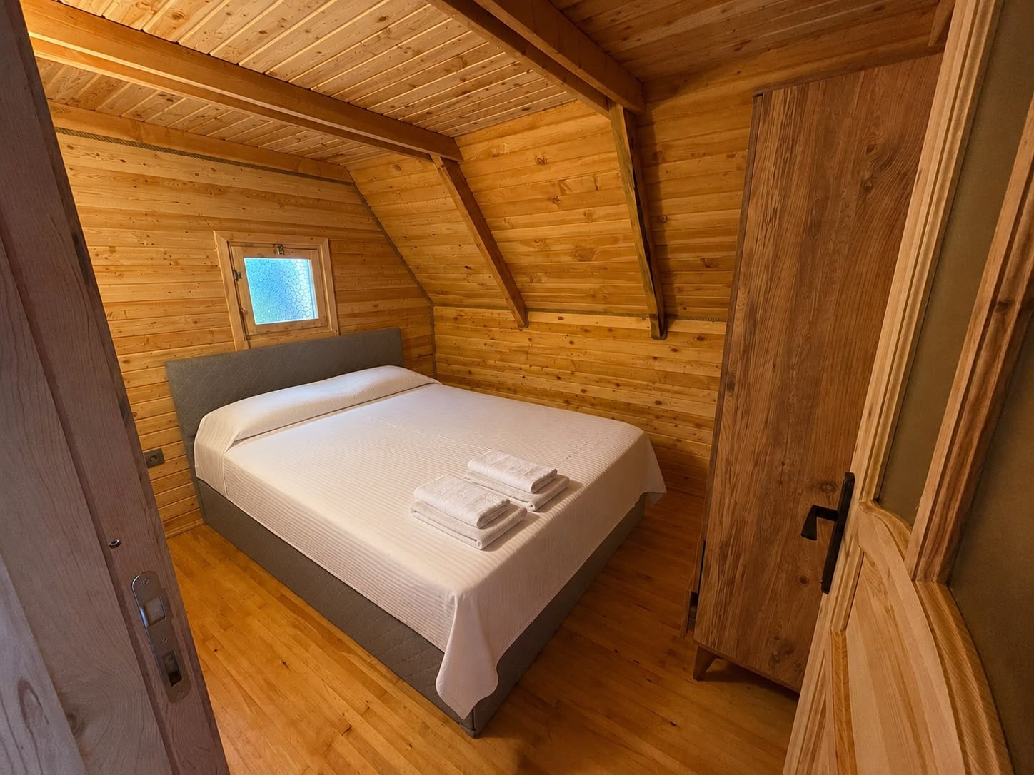 Bedroom with a made double bed, folded towels, timber walls and a small window.